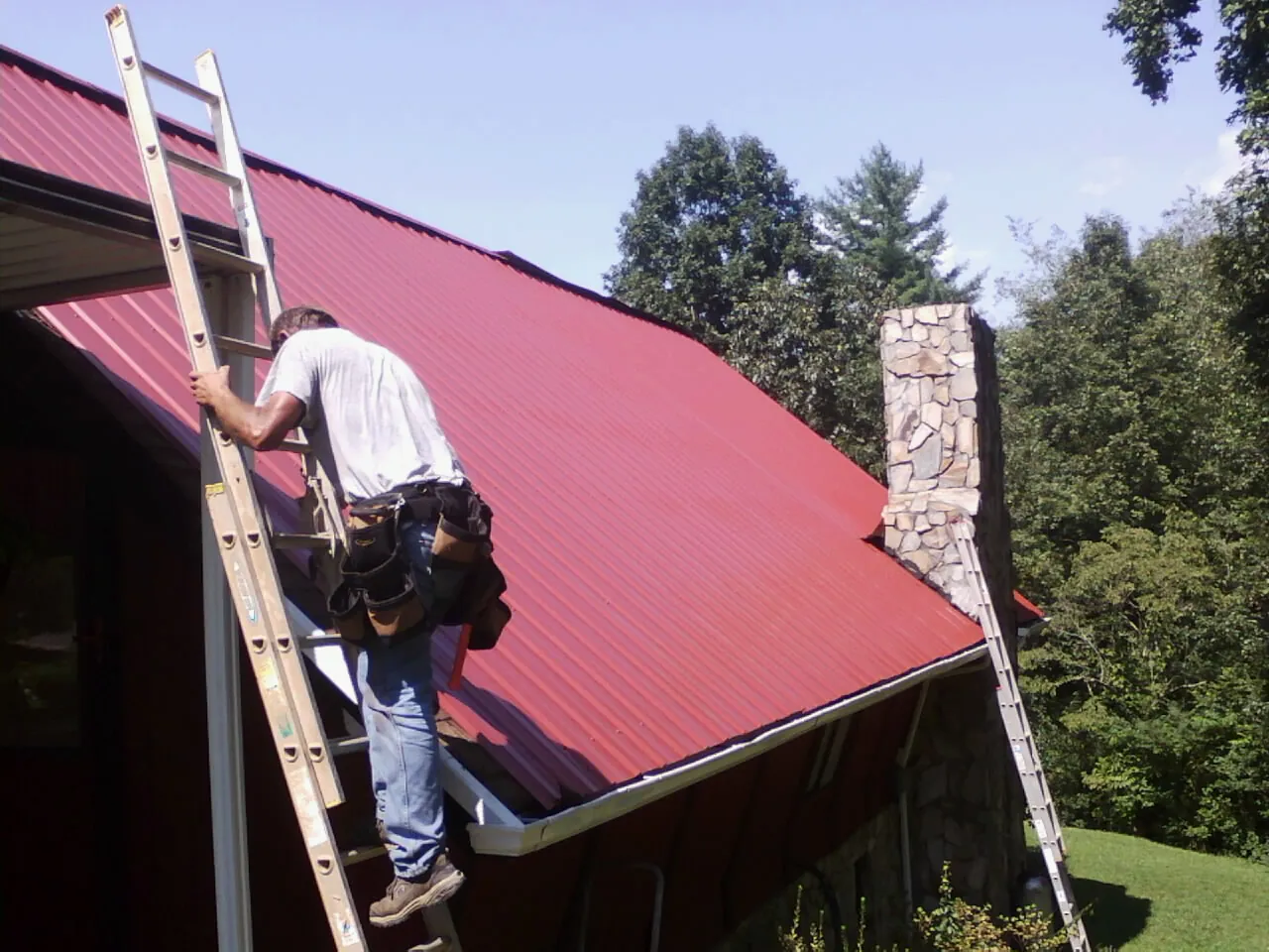 Roofer on a ladder beside a red metal roof and stone chimney — Soffit & Fascia Repair by Roofing Brothers in Rivesville